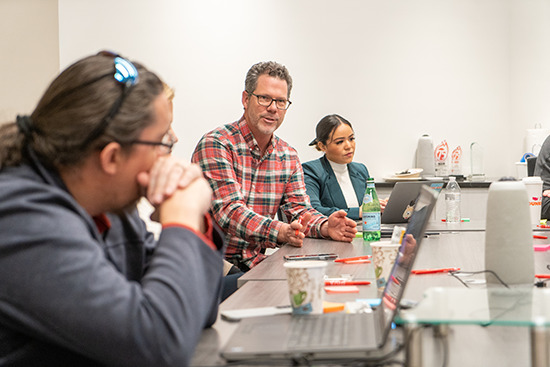 Man in red and grey plaid shirt deep in discussion with colleagues in conference room. Laptops and multi-colored sticky notes are on the conference table.